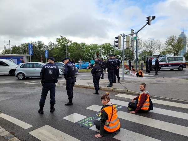 Man sieht einen Verkehrskreuzung, mit verschiedenen Autos. Im Vordergrund sitzen zwei Aktivist:innen der Letzten Generation mit orangen Warnwesten am Boden auf einem Zebrastreifen und schauen direkt in die Kamera. Vor ihnen liegt ein schwarzes Banner mit grüner, gelber und weißer Schrift. Vor ihnen stehen mehrere Polizist:innen mit dem Rücken zur Kamera. Weiter im Hintergrund sitzen andere Aktivist:innen ebenfalls auf einem Zebrastreifen, hinter ihnen stehen weitere Polizist:innen und ein großes Polizeiauto