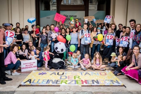 Das Foto zeigt eine große Gruppe von Menschen, die in die Kamera schauen. Sie sind bunt angezogen und halten Schilder gegen den 12-Stunden-Tag hoch. Vor den Menschen liegt ein großes Transparent mit der Aufschrift: "Aufstand der Alleinerziehenden".