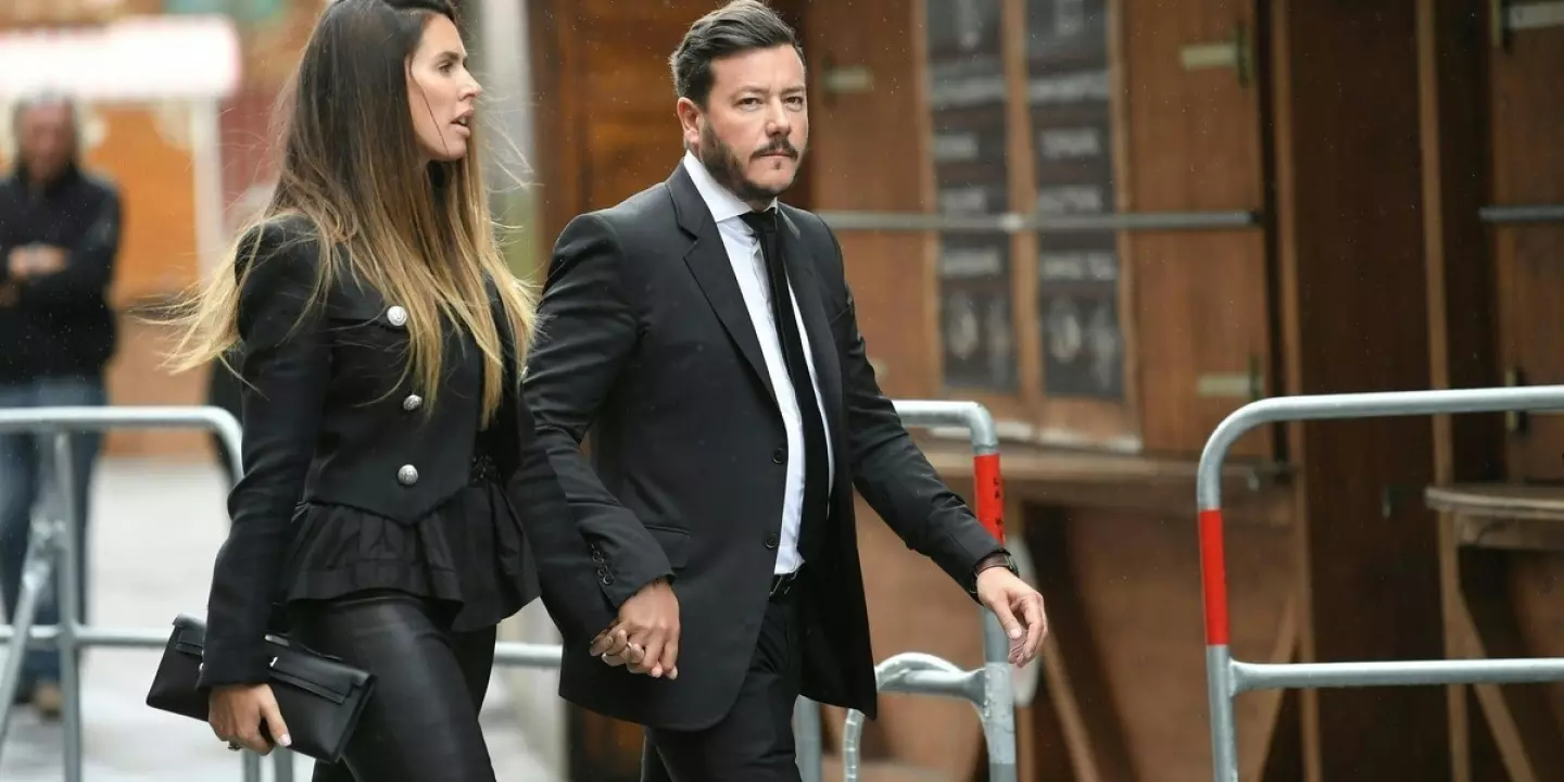 Man sieht René Benko im schwarzen Anzug mit, er geht händchenhaltend mit seiner Frau eine Straße vor einem Gebäude und Absperrgittern entlang.
