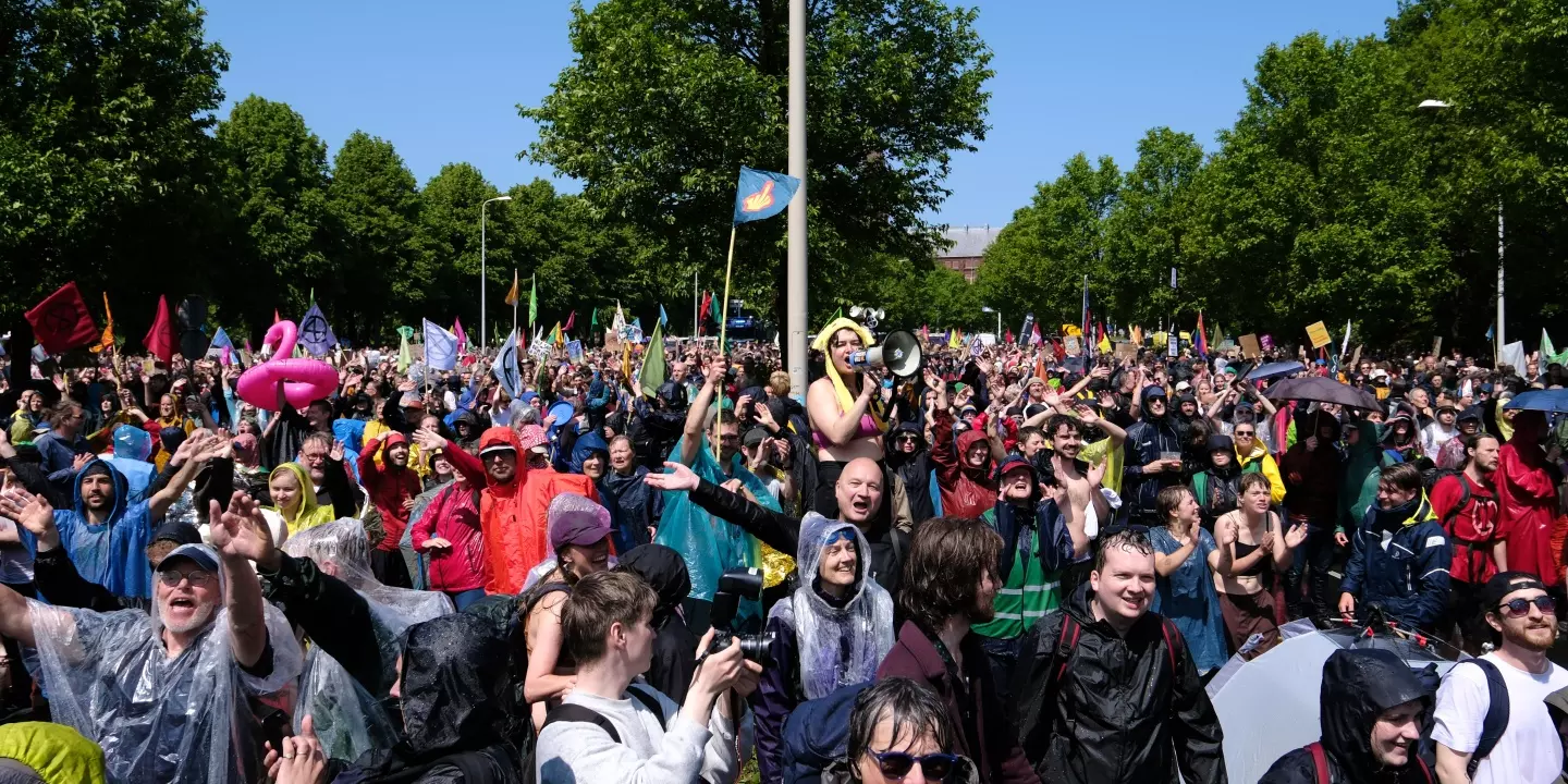 Eine Demonstration mit vielen Menschen, die nass aber fröhlich aussehen. Im Hintergrund grüne Bäume und blauer Himmel.