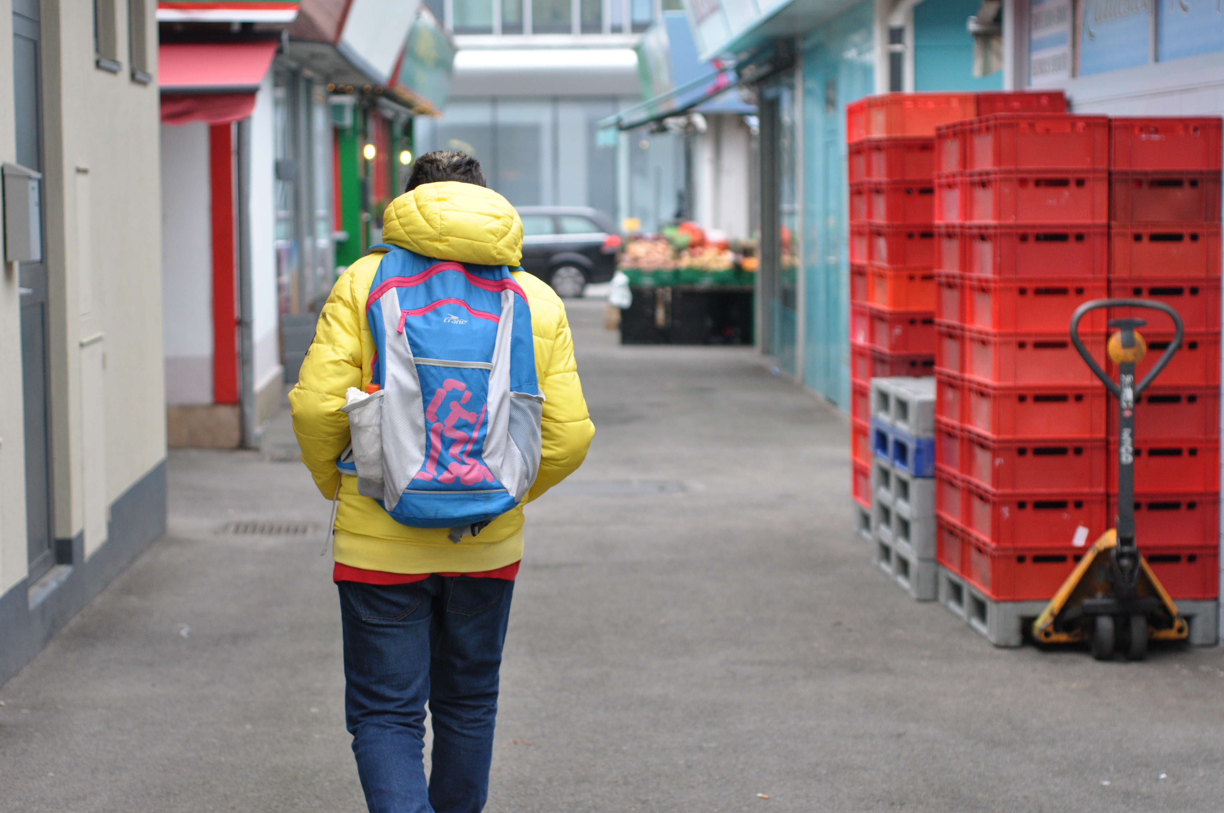 Soufiene mit Schultasche von hinten, wie er durch einen Markt geht. Die Hände hat er in die Taschen seiner gelben Jacke gesteckt.