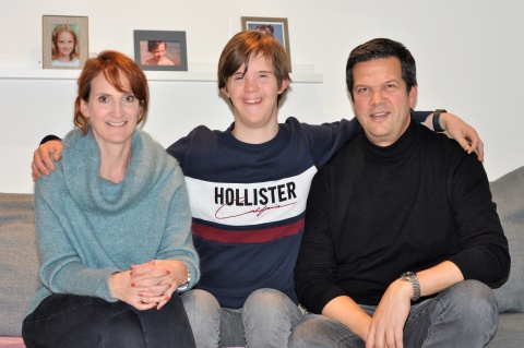 Claudia, Julian und Bernd Mühlbacher auf einem Sofa. Julian hat die Arme um seine Eltern gelegt, im Hintergrund Fotos seiner Schwester.