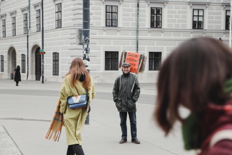 "Petar Thunberg" steht mit seinem Schild vor dem Bundeskanzleramt. Zwei Frauen gehen an ihm vorbei. Er lächelt.