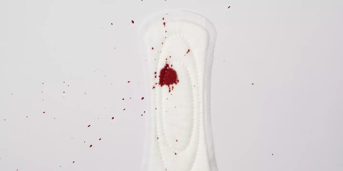 Ein weiße Menstruationsbinde liegt auf weißem Hintergrund. Von oben fällt ein Blutklecks darauf.