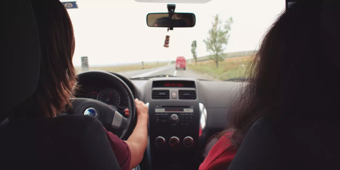 Von der Rückbank werden zwei Menschen in einem Auto fotografiert. Einer sitzt auf dem Fahrer-, einer auf dem Beifahrersitz. Durch die Winschutzscheibe sieht man noch ein Auto auf der Straße.