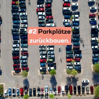 Eine Vogelaufnahme von einem Parkplatz. Es stehen unzählige Autos nah aneinander, dazwischen sind ein paar Menschen zu erkennen. “#2 Parkplätze zurückbauen”