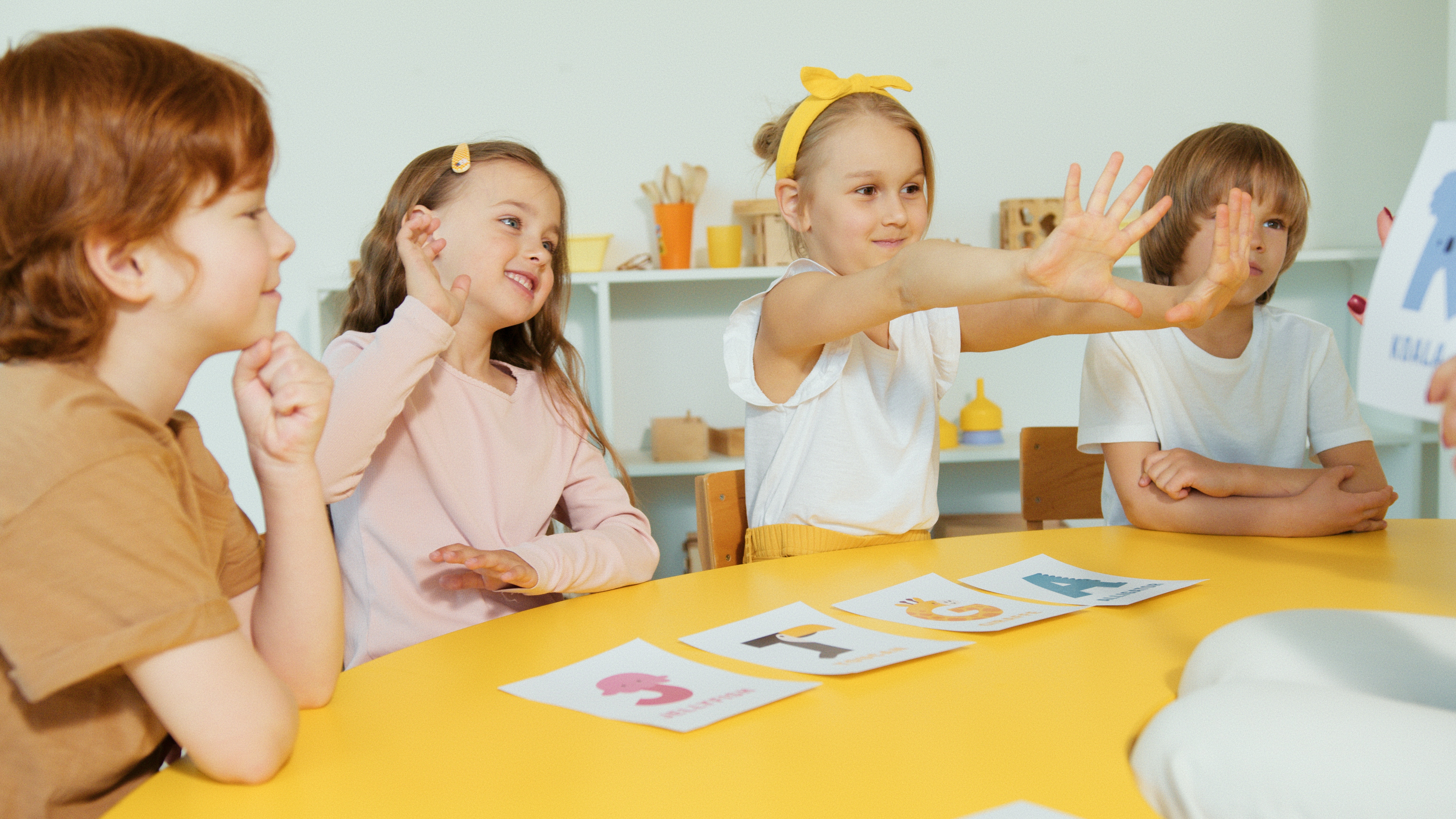 Kindergartenkinder sitzen um einen Tisch und zeigen mit ihren Händen nach vorne.