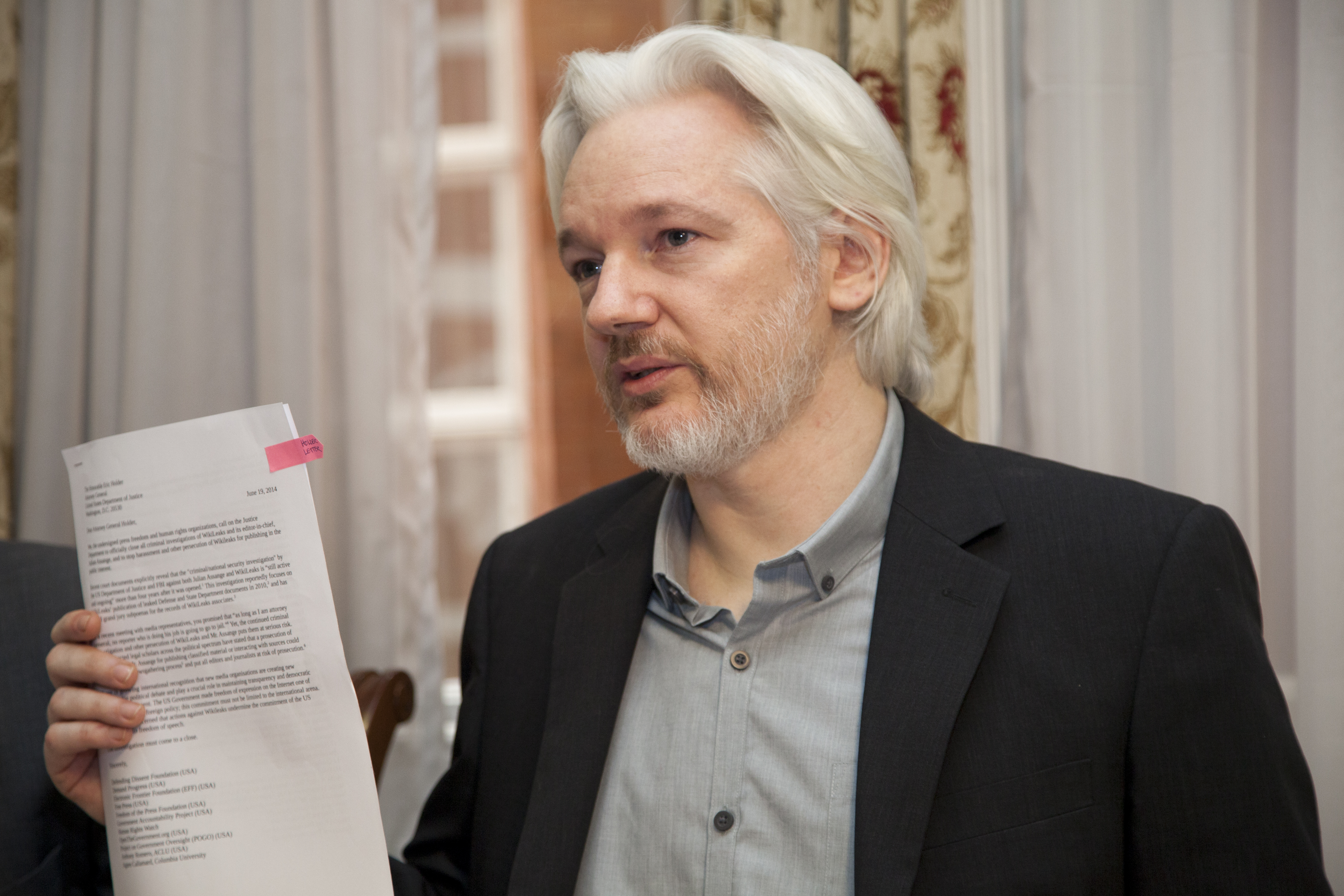 Julian Assange steht in der ecuadorianischen Botschaft in London und zeigt ein Dokument in die Kamera. Er hat lange weiße Haare und einen Bart