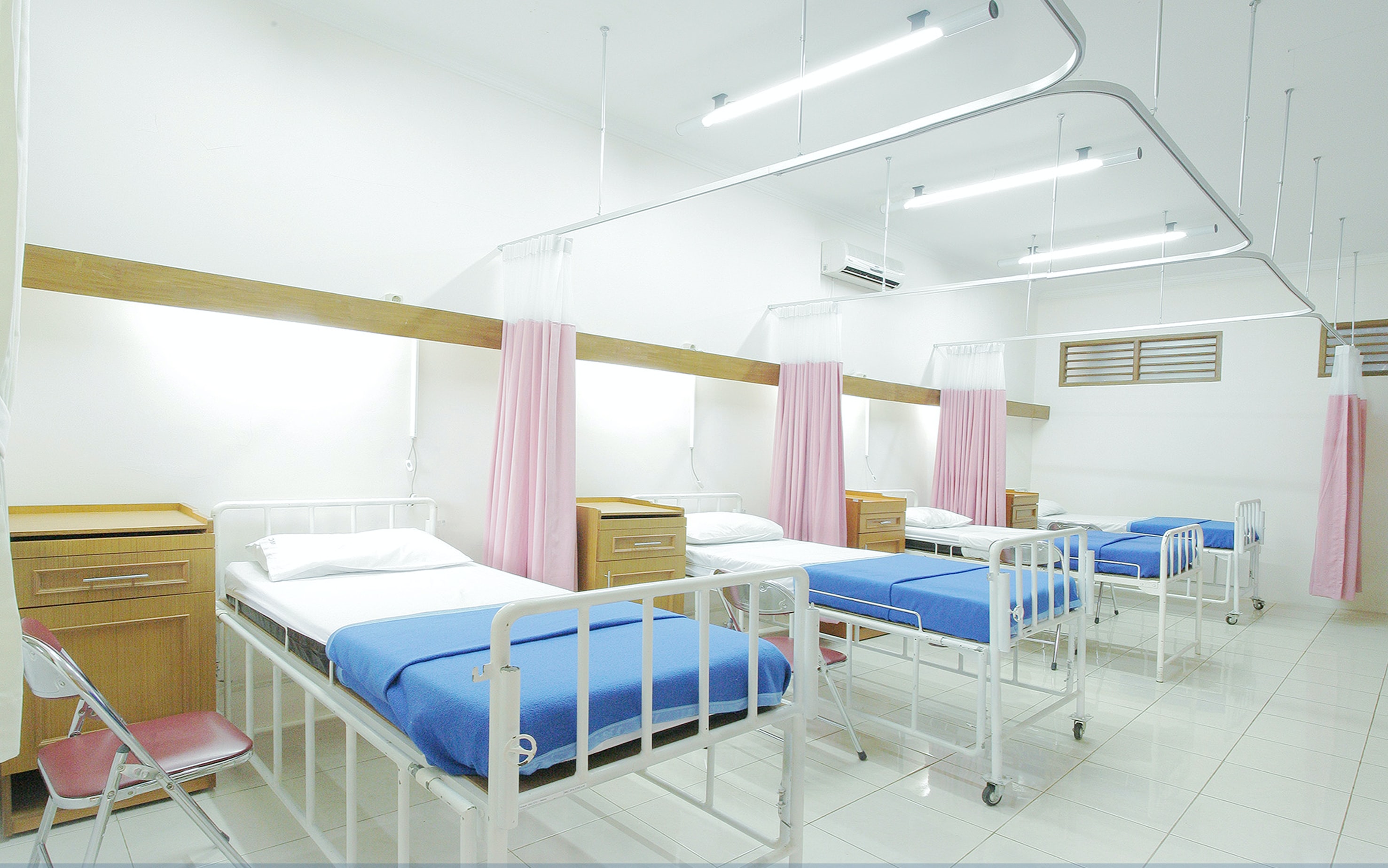 Drei Spitalsbetten mit blauen Decken und weißen Pölstern und rosa Vorhängen zwischen den Betten