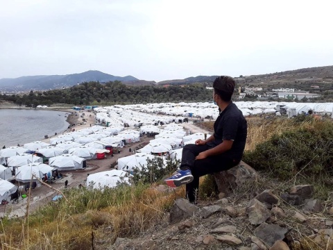 Ein Mann, in schwarz gekleidet, sitzt auf einem Hügel, unter ihm das Lager Kara Tepe