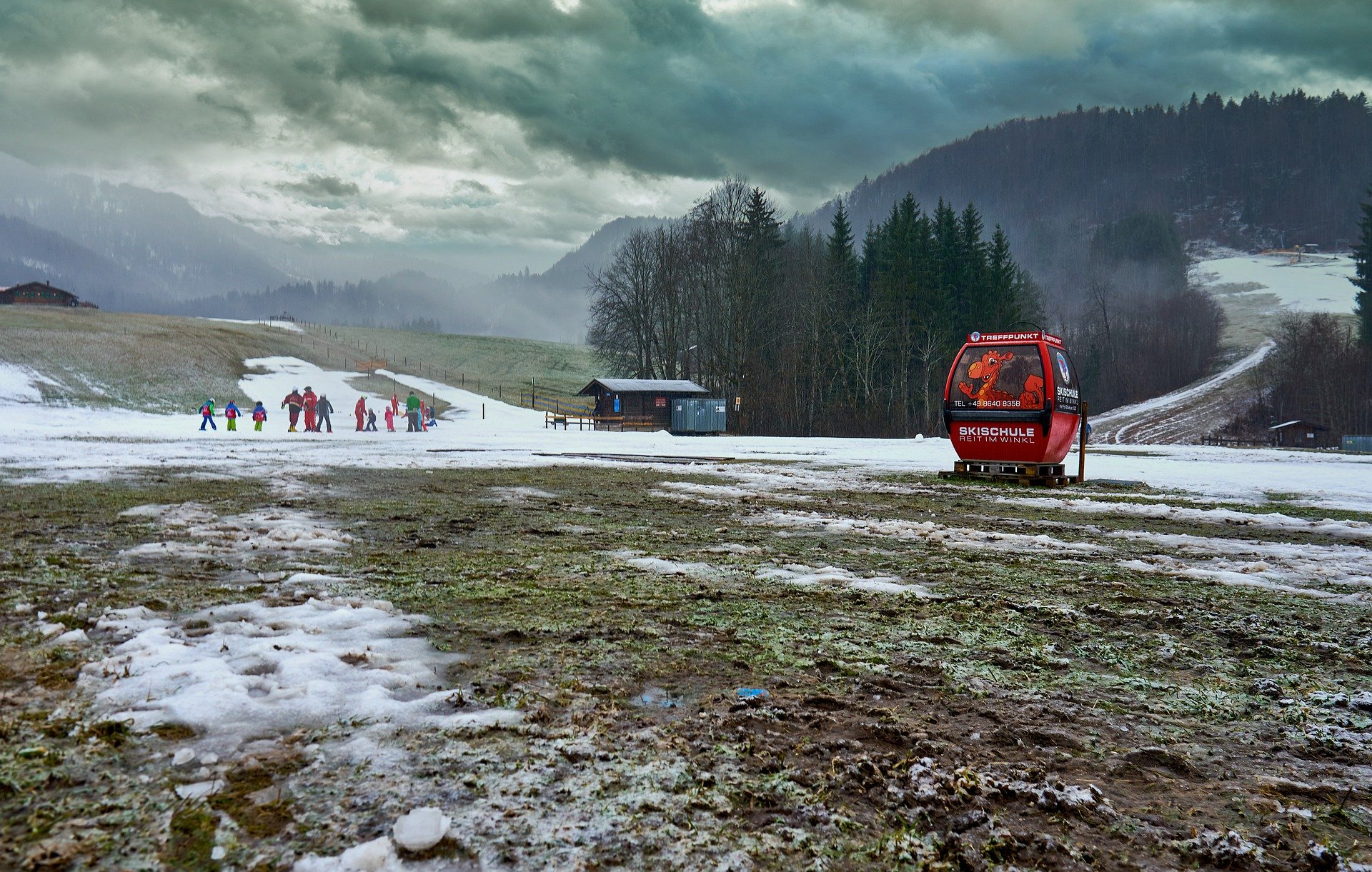 Bild einer Skigondel auf einem spärlich mit Schnee bedeckten Feld. Im Hintergrund ist eine Skischulgruppe zu sehen, die sich auf dem wenigen Schnee fortbewegen.