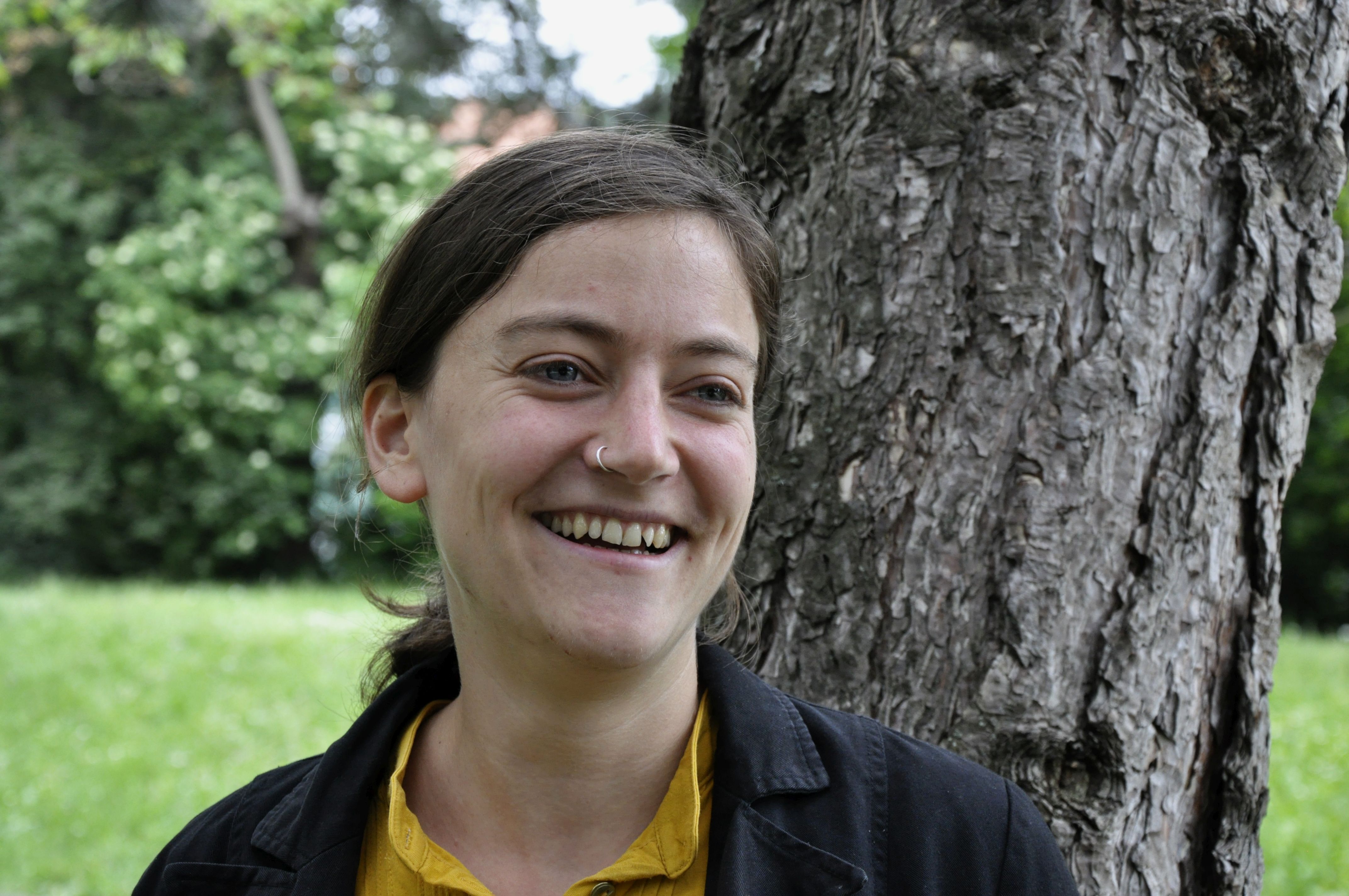 Sarah Zeller lächelt, einen Baum im Rücken, in die Kamera.