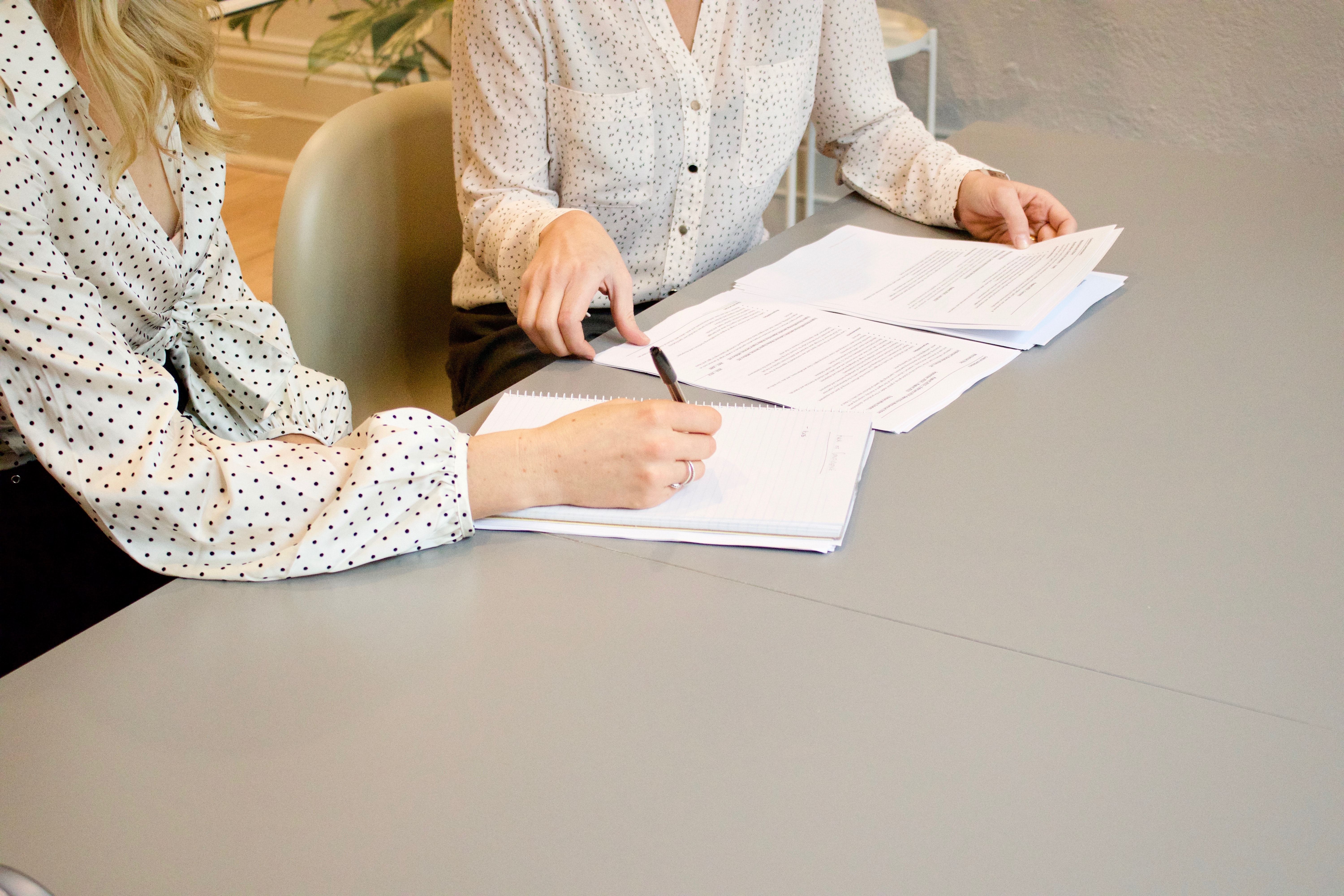 Oberkörper zwei weiblich gelesener Personen, die einander zugewandt sind, sind sichtbar. Beide tragen weiße Blusen und sitzen an einem Tisch auf dem Zettel liegen.
