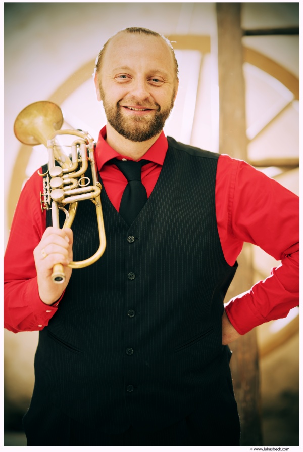 Der Trompeter Thomas Gansch trägt sein Instrument über die Schulter und lächelt in die Kamera.