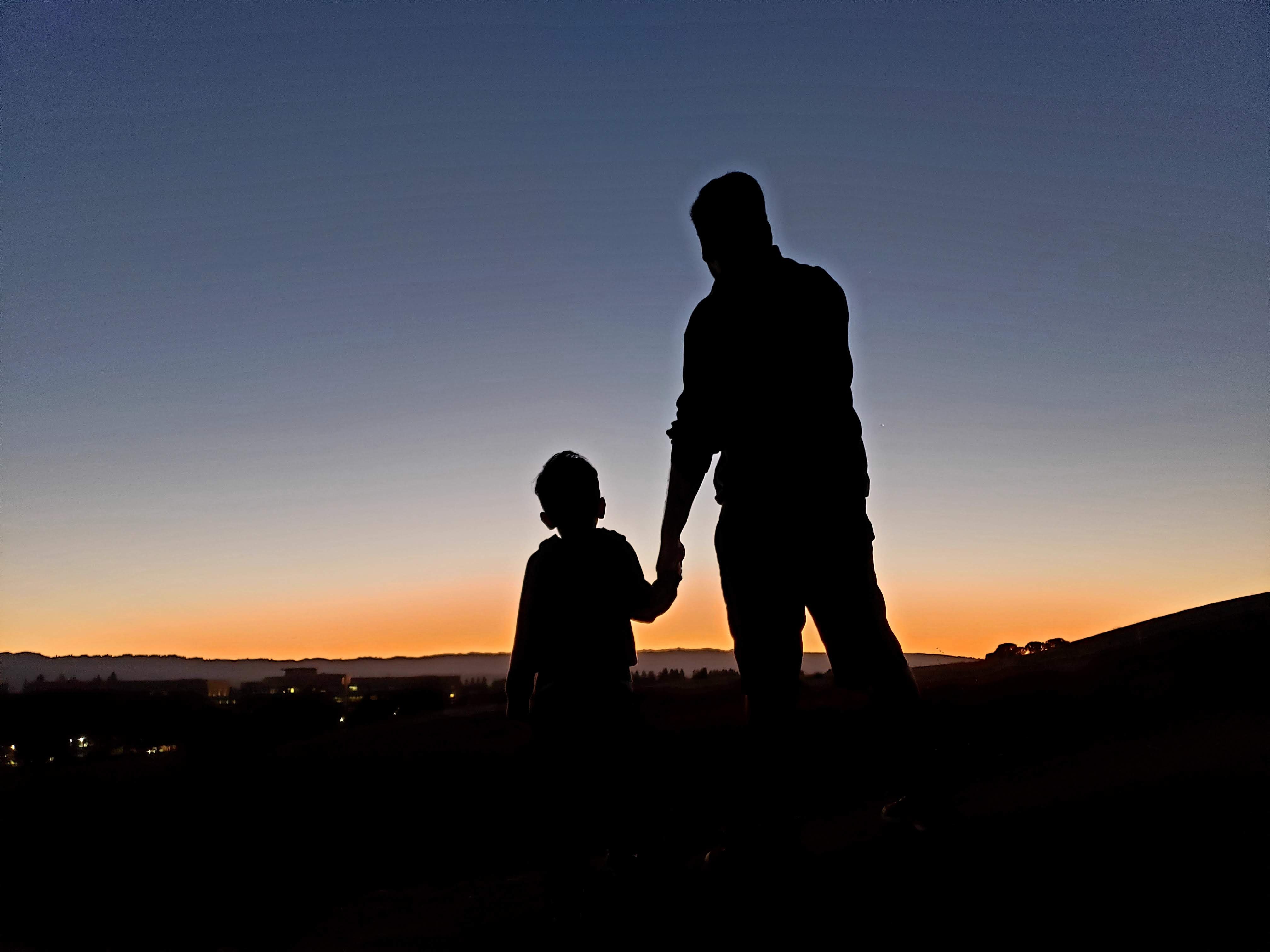 Ein Vater mit Depressionen erzählt, wie sich das anfühlt. Man sieht die Silhouetten einer erwachsenen Person, die ein Kind an einer Hand hält. Sie stehen vor einem Sonnenuntergang.