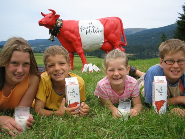 Kinder liegen auf einer Wiese und halten Milchgläser in die Kamera. Im Hintergrund steht eine rot-weiß-rote Kuh mit Faironika. Auf der Wiese liegen außerdem noch Packungen von "A faire Milch".