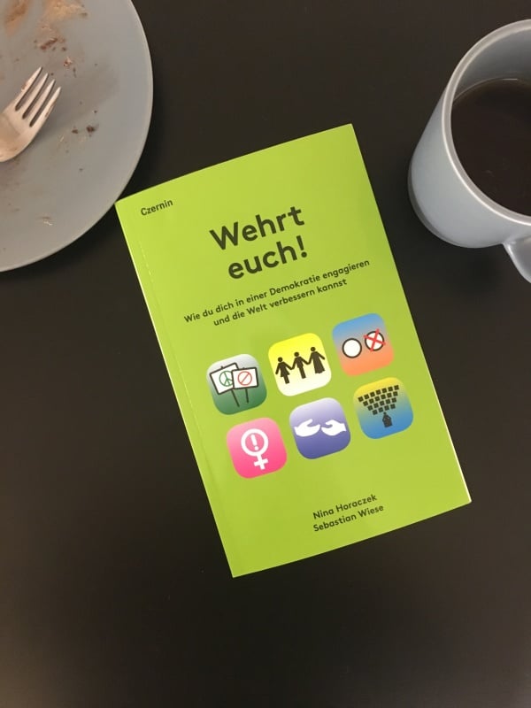 Ein Foto vom Buch "Wehrt euch" auf einem schwarzen Tisch mit Kaffeetasse und Kuchenteller