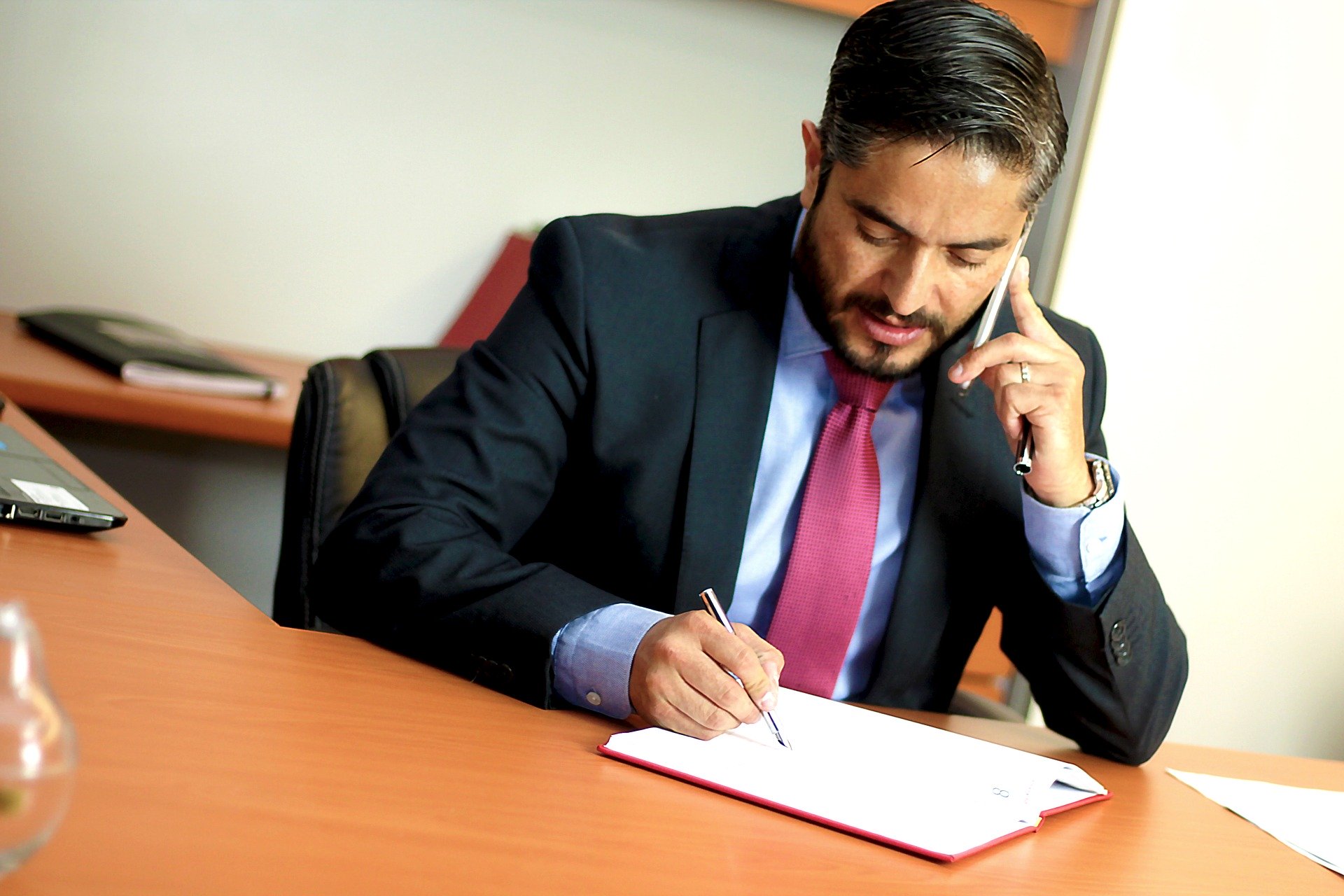 Ein Mann sitzt telefonierend an einem Schreibtisch. Er trägt einen Anzug und macht sich Notizen. Das Bild ist für einen Beitrag über die Arbeitsbedingungen von Rechtsanwaltsanwärter:innen.