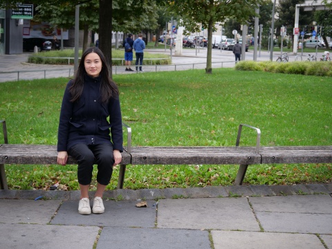 Virginia Lui sitzt auf einer Holzbank ohne Rückenlehne. Sie ist komplett schwarz gekleidet und sieht direkt in die Kamera.