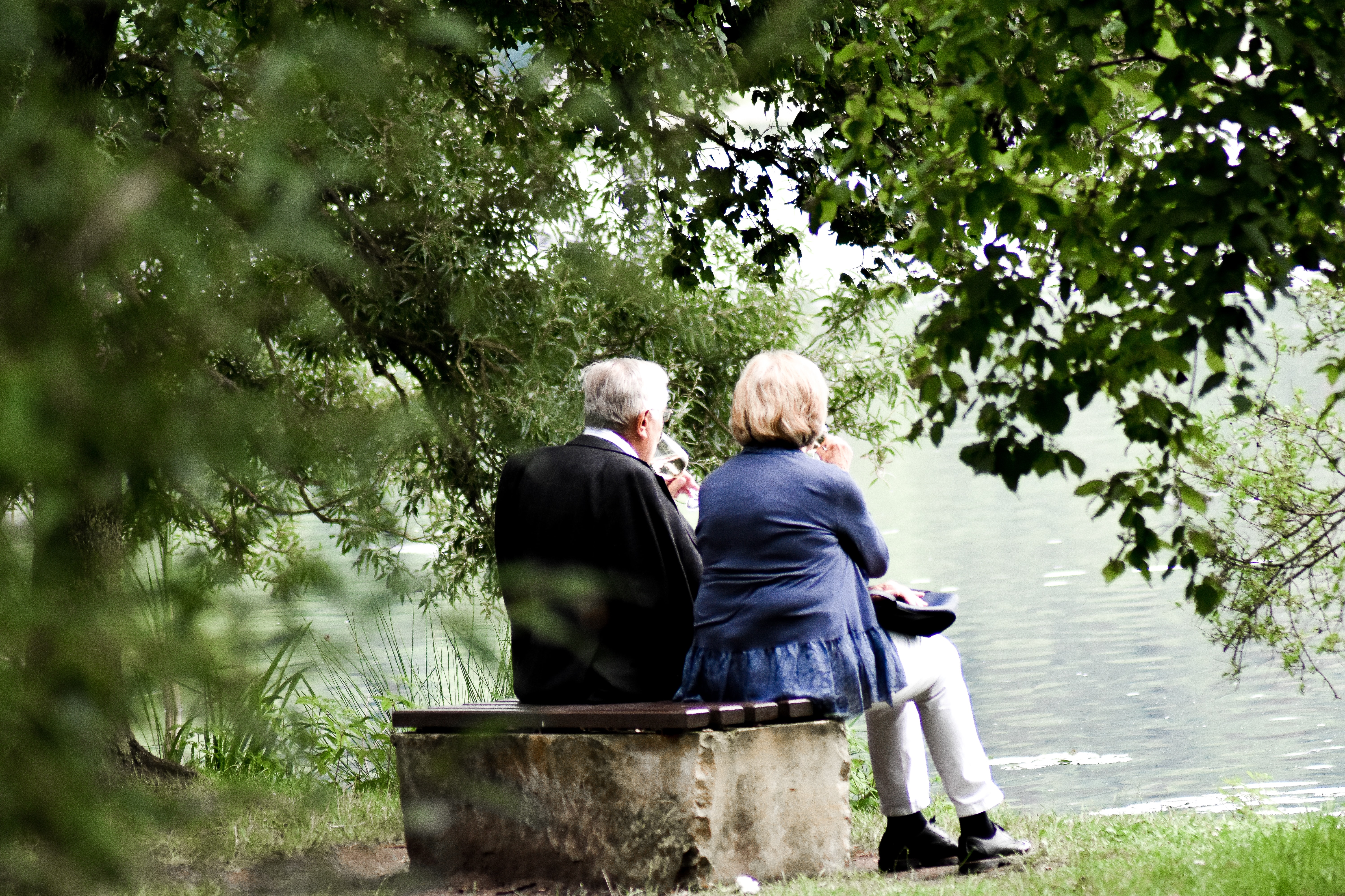 Foto zeigt ein älteres Paar an See, beide haben ein Glas Weißwein in der Hand und nippen an dem Getränk während sie auf den See blicken.