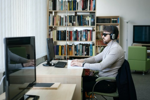 Symboldbild für Digitalisierung und Barrierefreiheit: Ein offenbar blinder Mann sitzt vor einem PC-Bildschirm. Er liest mit seinen Händen etwas von einem Blatt Papier ab.