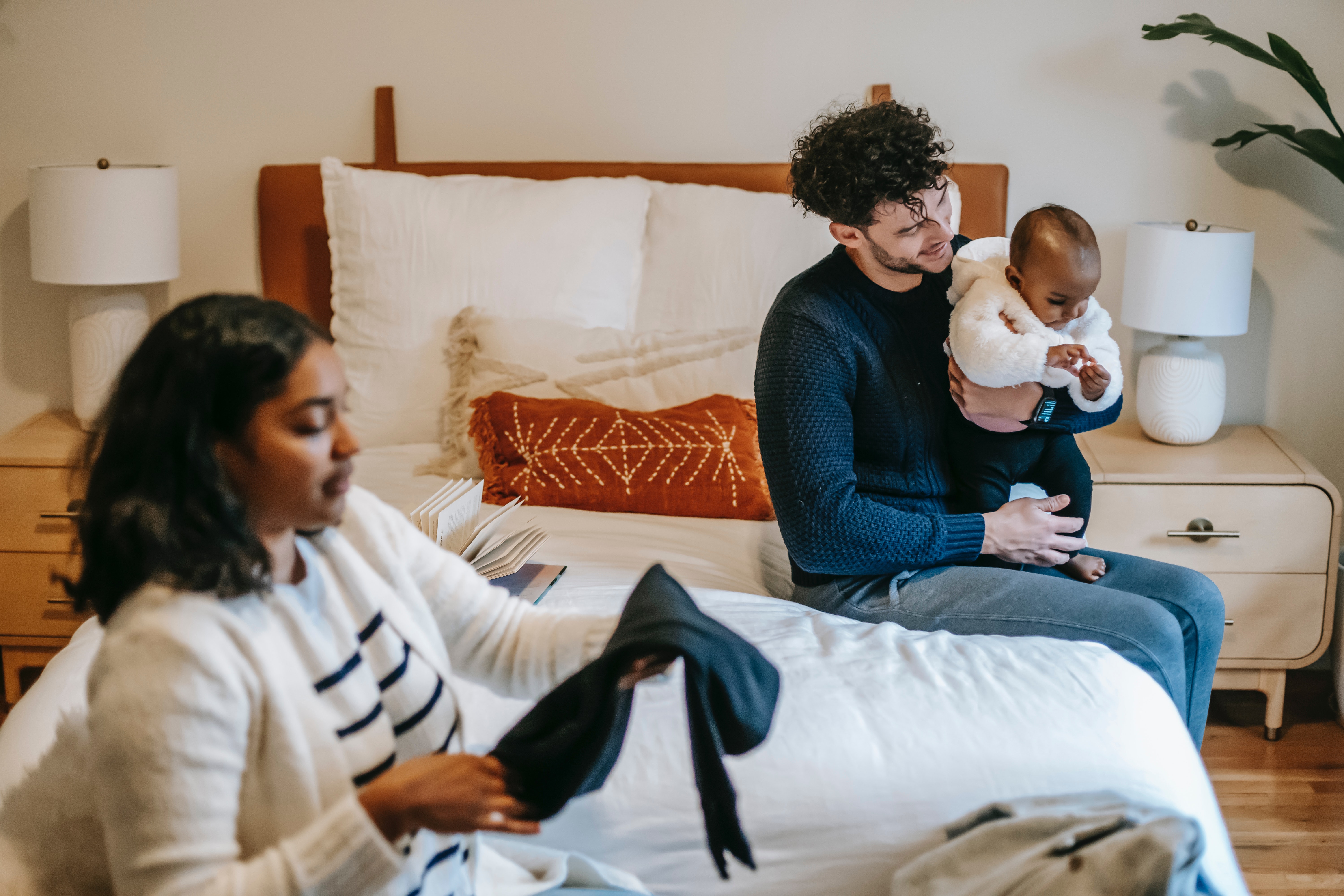 Eine Frau und ein Mann sind zu sehen. Sie sitzen auf einem Bett. Der Mann hält ein Kind im Arm. Bebildert wird damit ein Artikel zu Mental Load.