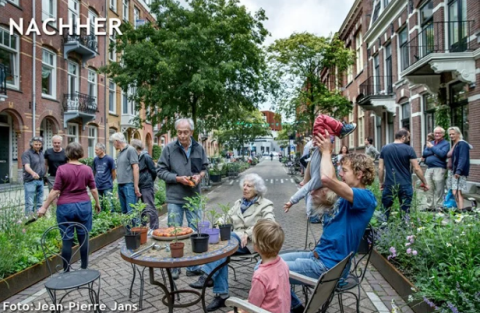 Das Bild zeigt die gleiche, nun von Autos befreite Straße, auf der statt Parkplätzen Blumenbeete stehen und Gruppen an Menschen auf der Straße sitzen.