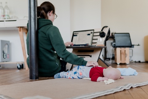 Eine Frau lehnt an einem Pfosten während sie auf dem Laptop arbeitet und mit der anderen Hand ihr Kleinkind streichelt.