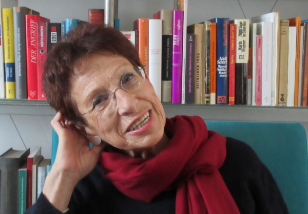 Ruth Wodak beschäftigt sich mit den Auswirkungen von Sprache. Sie sitzt vor einem Bücherregal und blickt lachend in die Kamera.