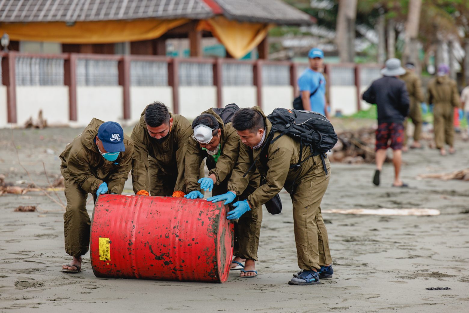 Menschen beheben eine Ölkatastrophe auf den Philipinnen. Mit Masken und Overalls geschützte Personen rollen ein Ölfass über einen verdreckten Strand. Foto: Project VIP