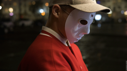 Ein Mann mit Kappe und roter Jacke vor dunklem Hintergrund. Er trägt eine Hockey-Maske im Gesicht