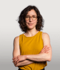 Foto von Ruth Eisenreich. Sie hat halblange wellige braune Haare und trägt eine Brille und ein gelbes Tanktop.