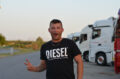 Rafal zeigt mit zwei ausgestreckten Fingern das Victory-Zeichen. Er steht vor aufgereihten Trucks im Abendlicht und lächelt breit. Er ist trotz mancher Schwierigkeiten gerne Fernfahrer.