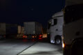 Nachts auf einer Raststätte. Die Scheinwerfer eines LKW beleuchten die Straße, ein anderer fährt gerade daran vorbei.