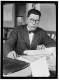 Schwarzweißfoto von Raymond Pearl. Er hat kurzes, schwarzes Haar, eine schwarze, runde Brille. Er trägt einen Anzug und eine dunkle Fliege. Er sitzt an einem Schreibtisch und hat einen strengen Blick.