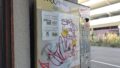 Das Foto zeigt einen Automaten, ähnlich einem Zigarettenautomaten, der an einer Wand hängt. Er ist mit Grafitti beschmiert, links hängt an ihm ein laminierter Zettel, auf dem unter dem Titel